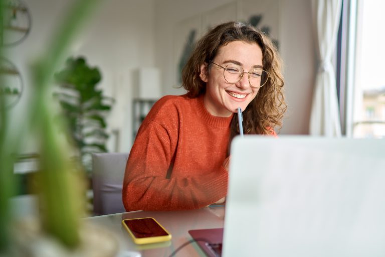 Ung glad kvinnlig student som använder en laptop, tittar på ett webbseminarium och antecknar hemma.